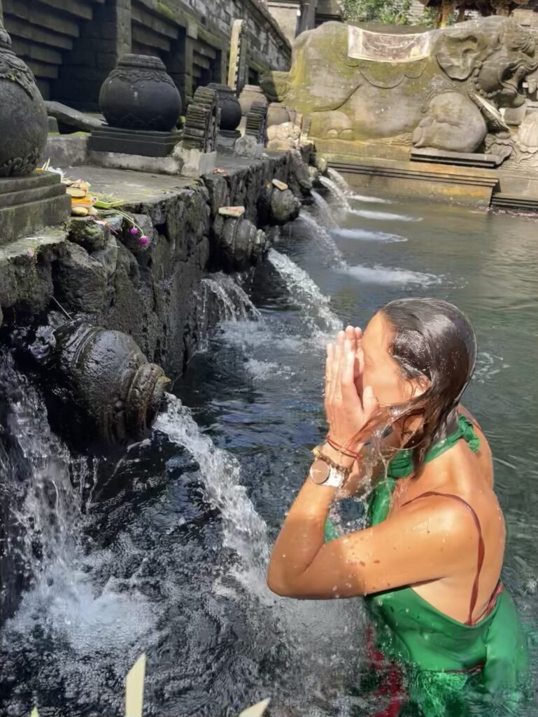 Céline participant à un rituel sacré au temple Pura Tirta Empul à Bali, aspergeant son visage avec de l’eau bénite dans un rituel de purification.