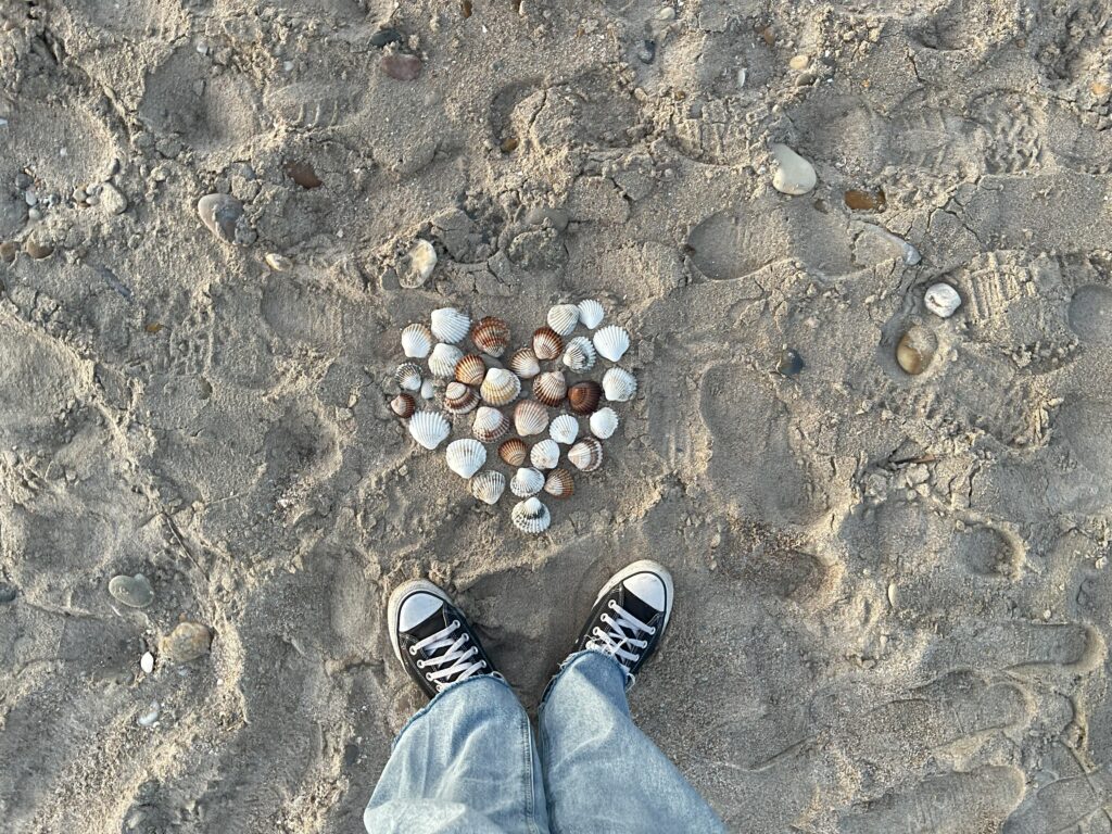 Deux pieds devant un cœur formé de coquillages sur le sable, illustrant l’histoire et la passion de Céline pour Immersive Expérience.