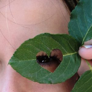 Femme regardant à travers une feuille en forme de cœur, symbolisant la reconnexion à soi offerte par le Massage Immersion Nidra : Rêve Éveillé. Ce soin combine le Yoga Nidra, la méditation, la respiration consciente et le massage pour une relaxation mentale et physique profonde.